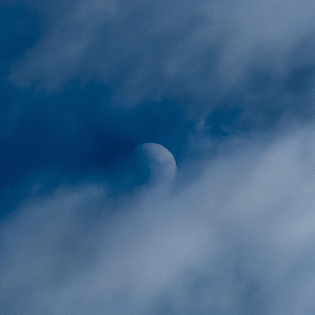 A moon barely visible through hazy clouds with a deep blue background